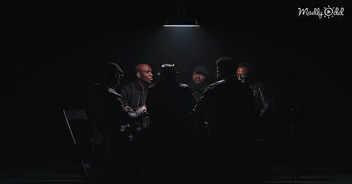 Six men in dark attire sit under a single overhead light in a dimly lit room.