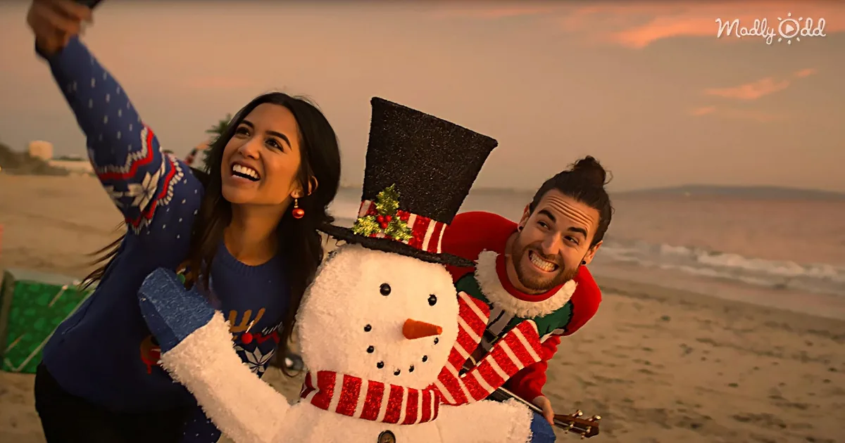 A woman in a festive sweater takes a selfie with a man in an elf costume and a snowman on a beach at sunset.