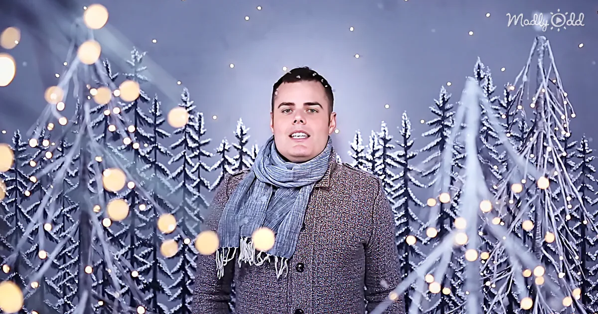 A man in a scarf and coat stands in front of a snowy forest backdrop with glowing lights.