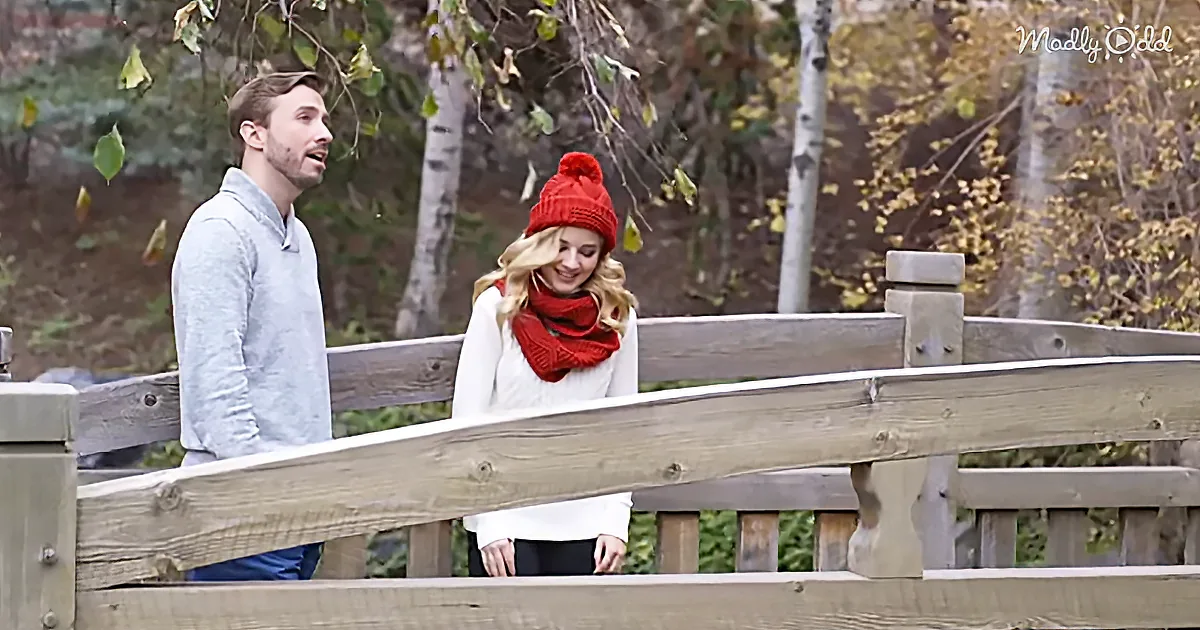 A man and woman stand on a wooden bridge, the woman wearing a red beanie and scarf, smiling.