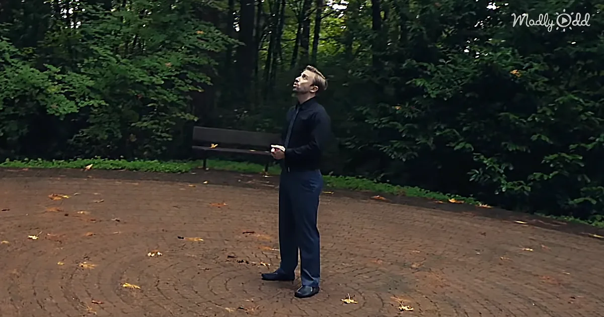 A man in a dark suit stands on a brick path, looking up, with a bench and greenery in the background.