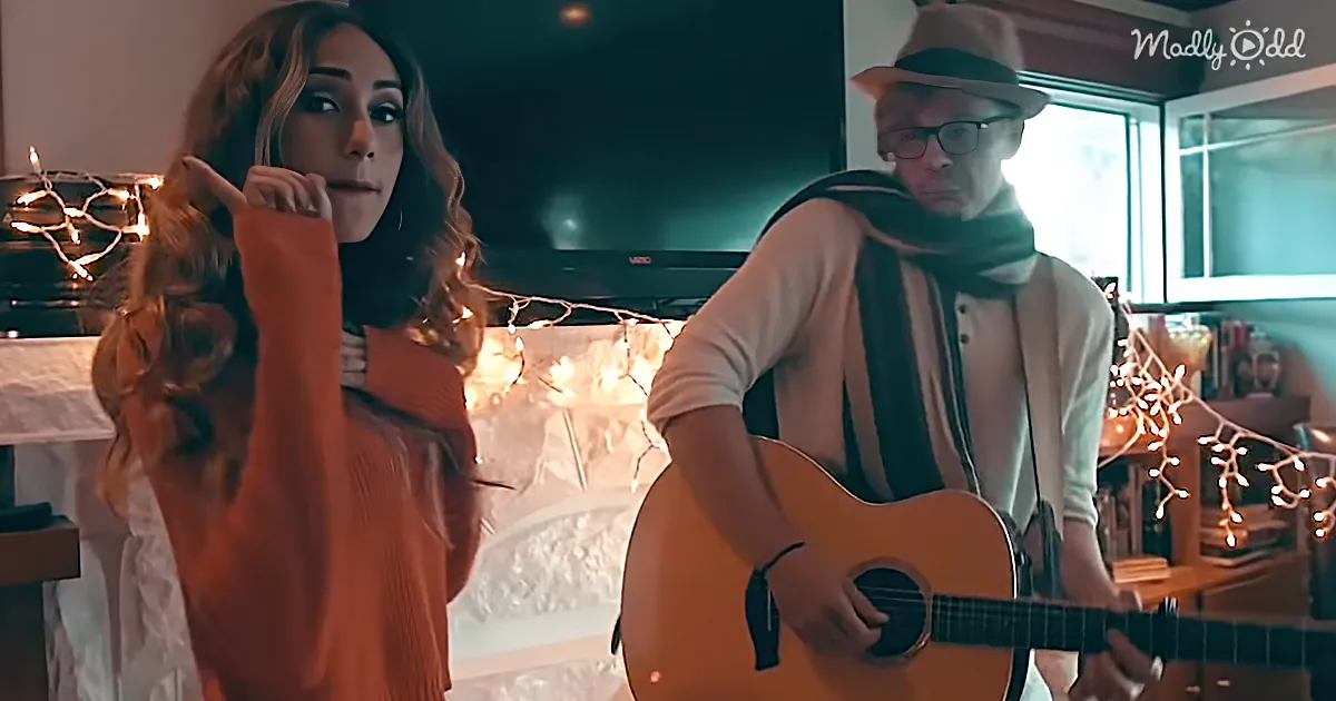 A woman in an orange sweater adjusts her hair while a man in a hat and scarf plays guitar in a cozy room with string lights.
