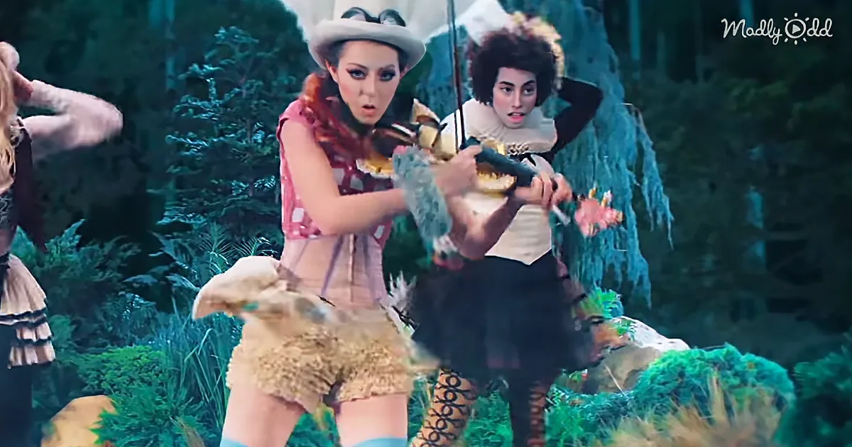 A woman in a whimsical costume plays a violin in a forest setting, surrounded by other performers.