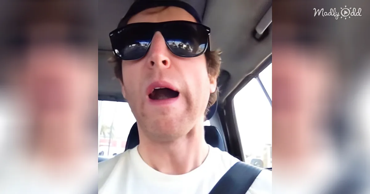 A person wearing sunglasses and a white shirt is inside a car, with a seatbelt on and a blurred background.