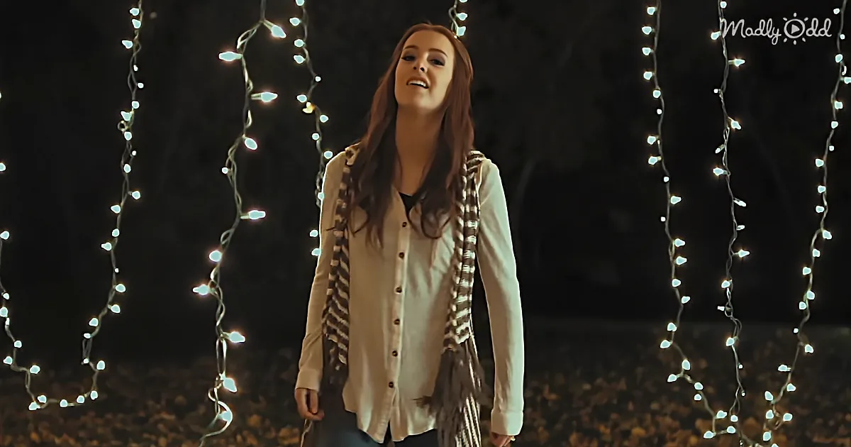 A woman stands amidst hanging string lights, wearing a white shirt and scarf, smiling upward.