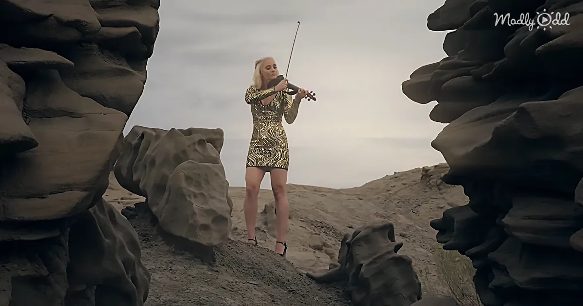 A woman in a gold dress plays violin amidst large, textured rock formations under a cloudy sky.