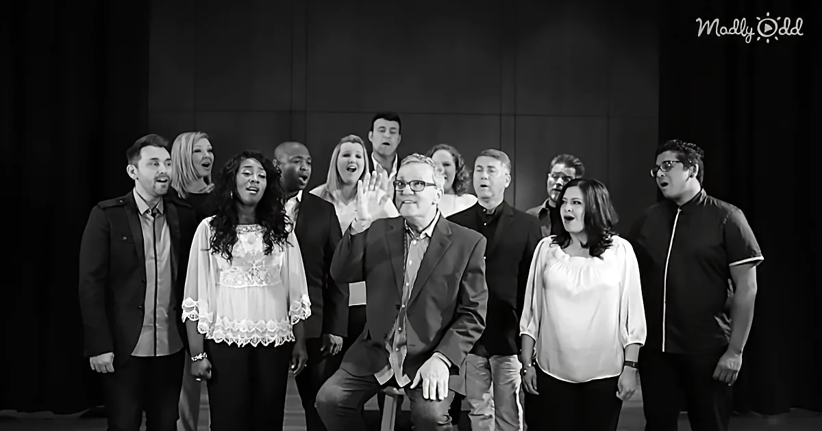 A man in a suit sits in front of a group of people singing, all in formal attire.