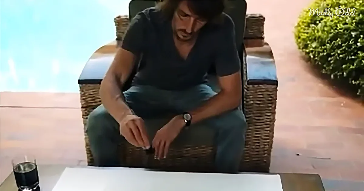 A man sits on a wicker chair outdoors, looking down at his hands, with a glass of dark liquid on a table nearby.