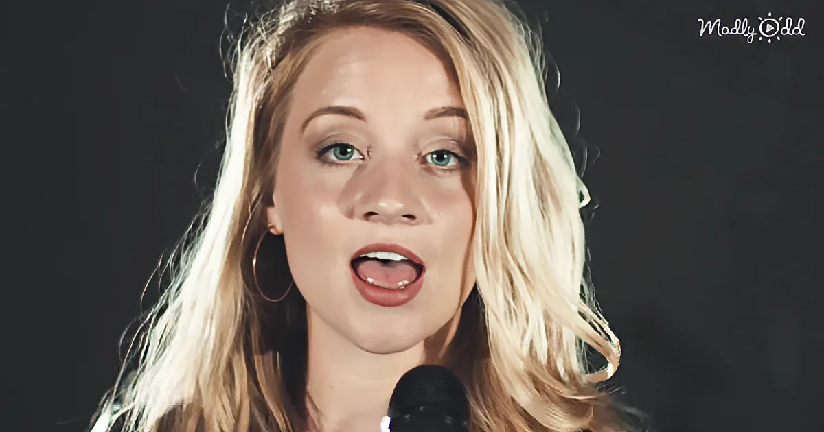 A person with long blonde hair and hoop earrings sings into a microphone against a dark background.