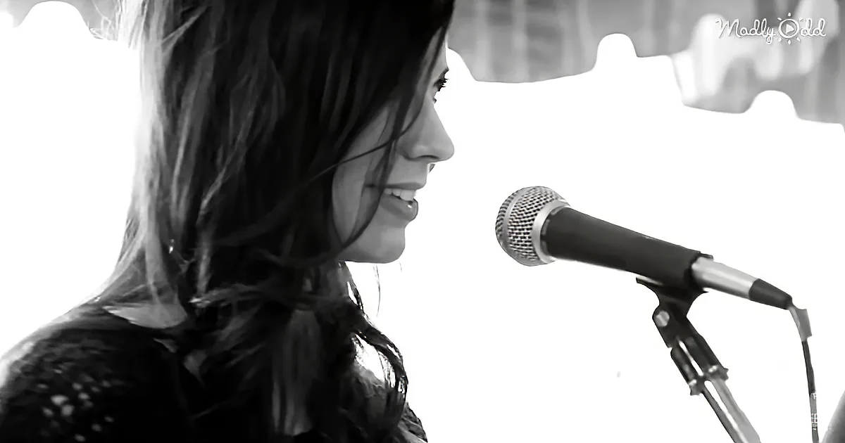 A woman with long hair smiles near a microphone, possibly singing or speaking.