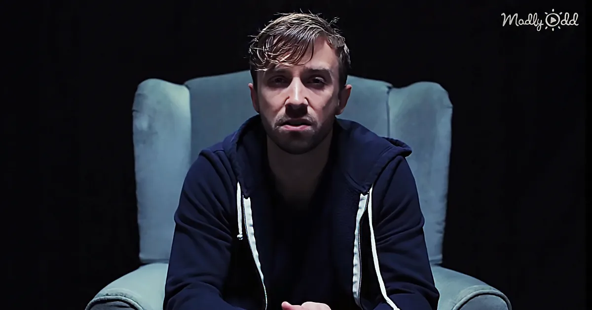 A man in a dark hoodie sits in a blue chair against a dark background.