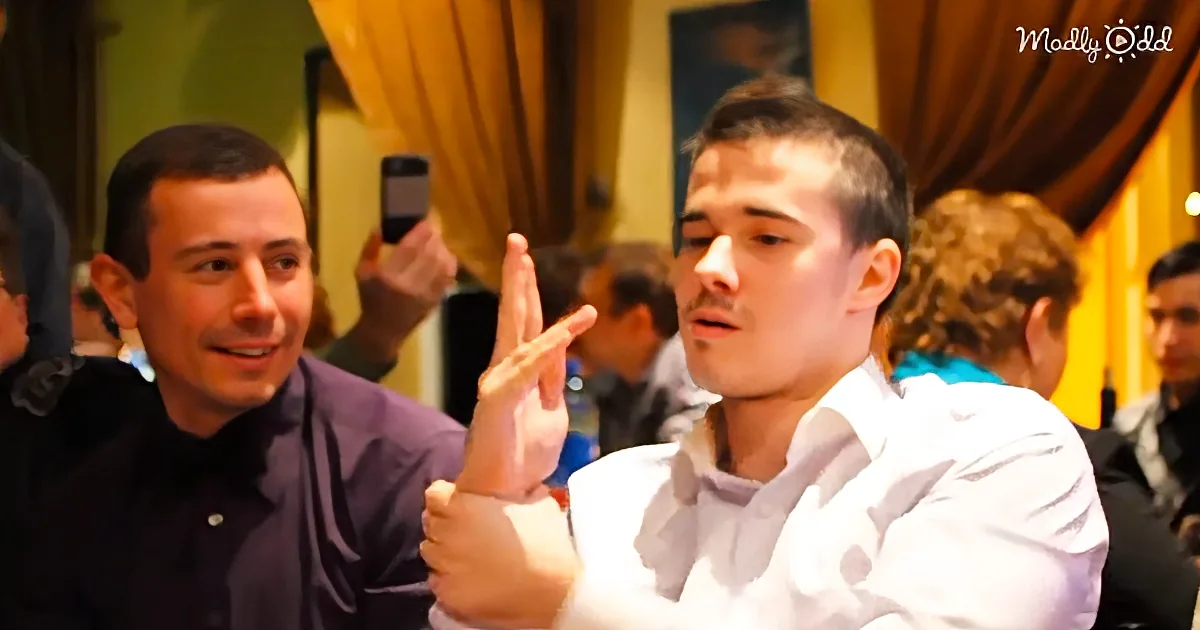 A man in a white shirt gestures with his hand while another man in a purple shirt smiles and holds up a phone.