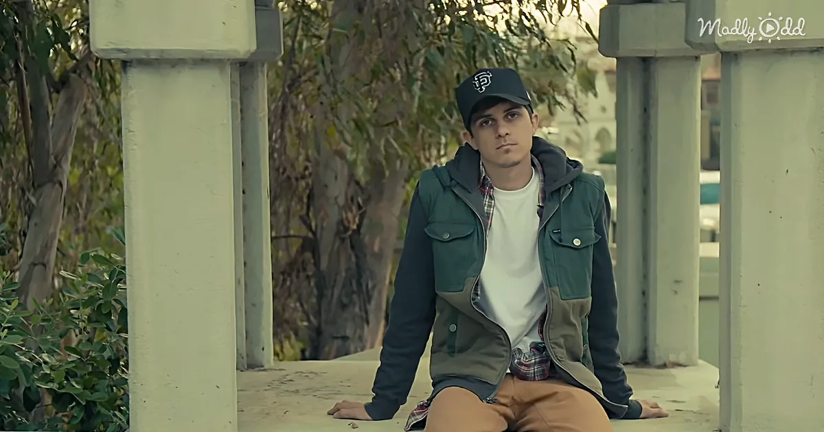 A person wearing a cap, jacket, and plaid shirt sits on a stone surface amidst columns and greenery.