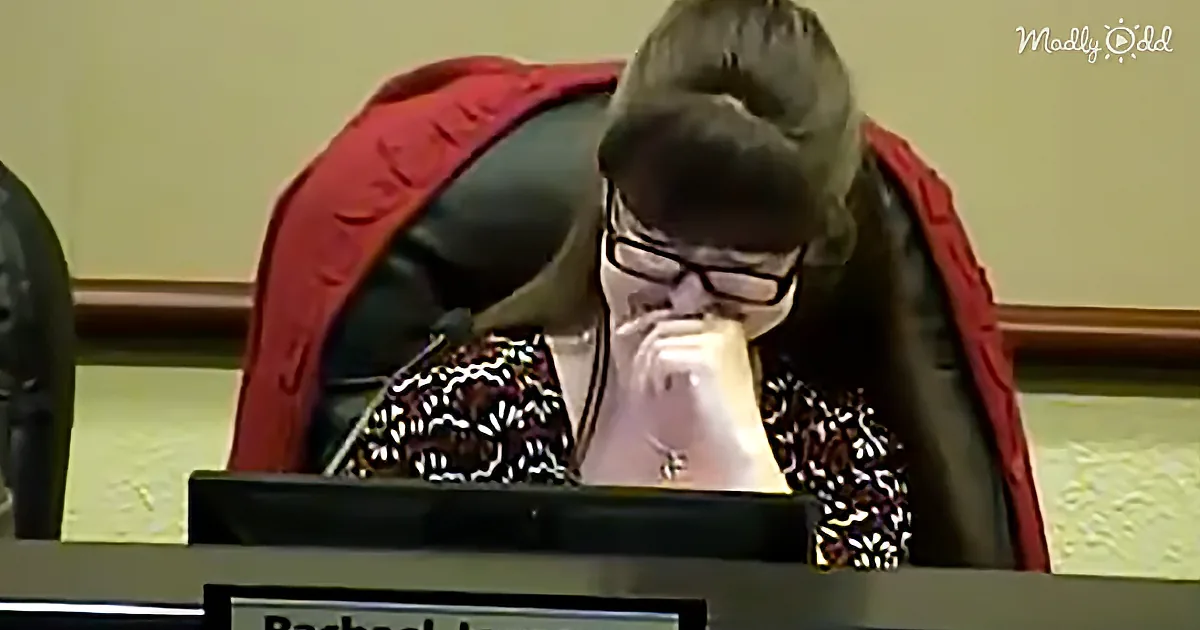 A woman with glasses sits at a desk, looking down, wearing a patterned top and a red jacket.