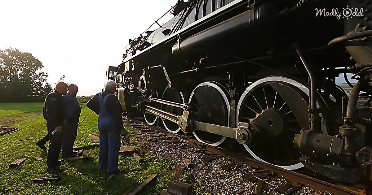 A group of people stands near a large steam locomotive on a railway track, examining its wheels and components.