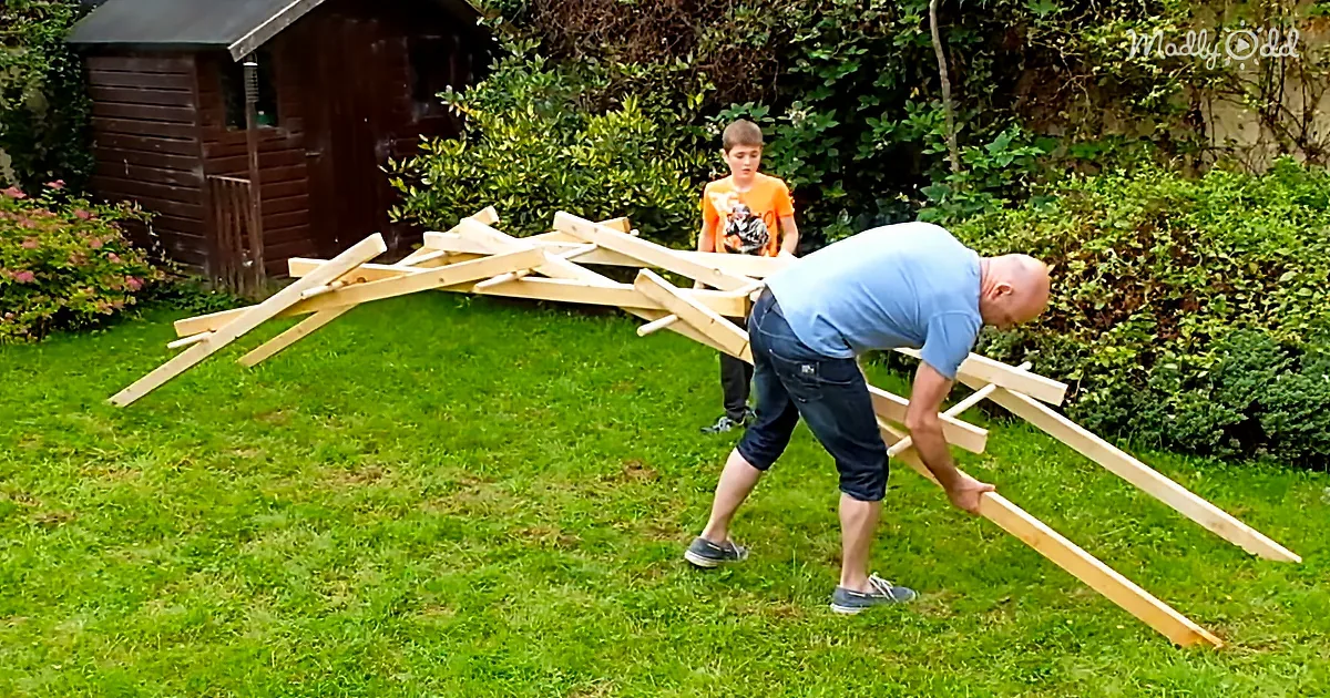 A man bends over a wooden structure on grass, with a boy in the background near a shed.