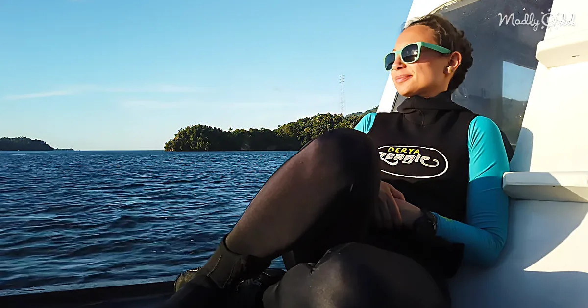 A woman in a wetsuit sits on a boat, wearing sunglasses, with a scenic view of the ocean and islands in the background.