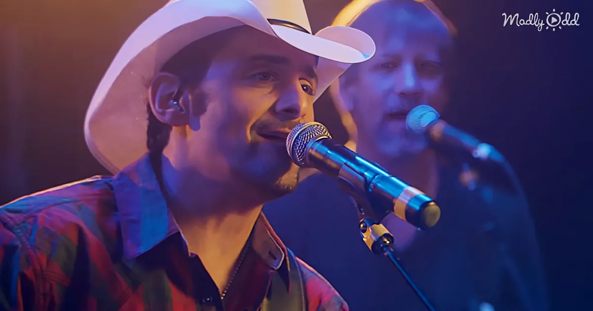 A man in a white cowboy hat sings into a microphone, wearing a plaid shirt, with another person in the background.