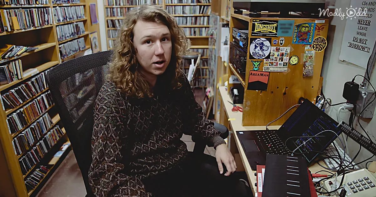 A person with curly hair sits in a chair, surrounded by shelves of CDs and a desk with a laptop and various equipment.