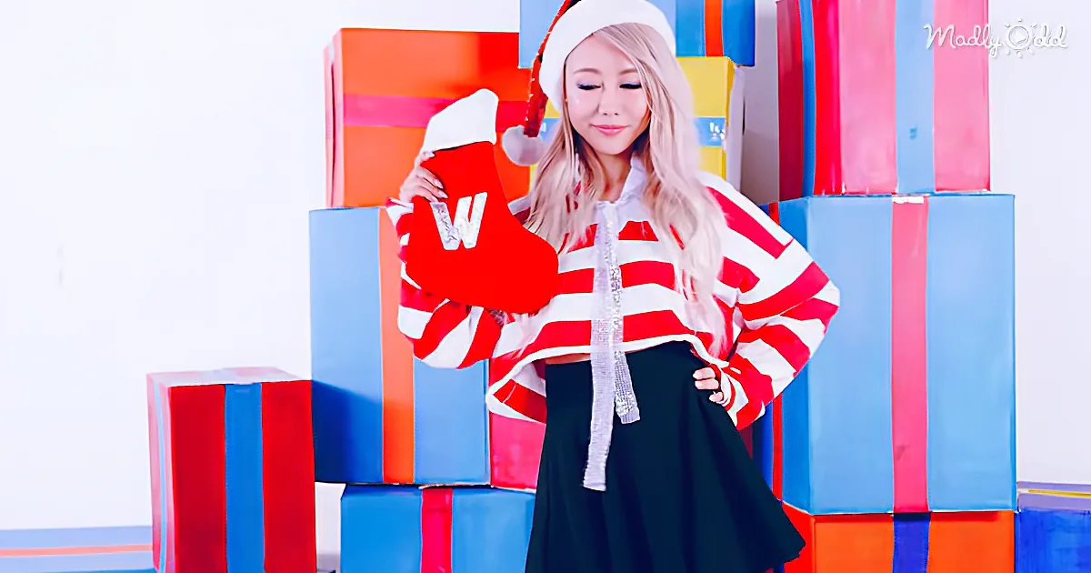 A person in a red and white striped sweater and Santa hat holds a red stocking, standing in front of colorful gift boxes.
