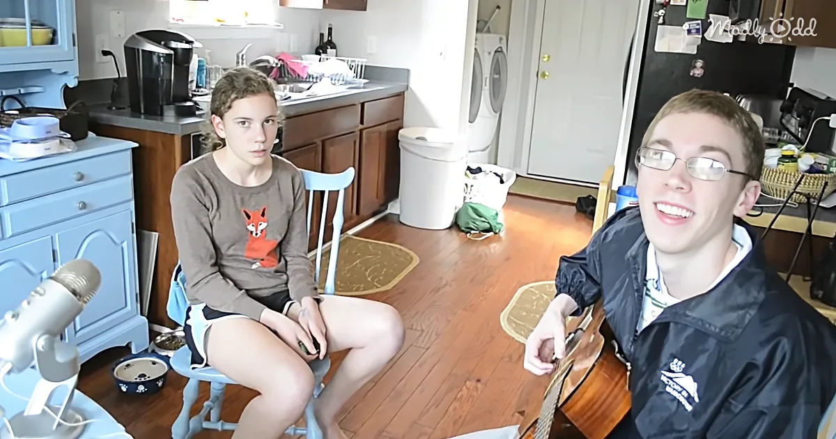 A young girl sits on a stool in a kitchen, while a boy in a jacket sits nearby, both facing the camera.
