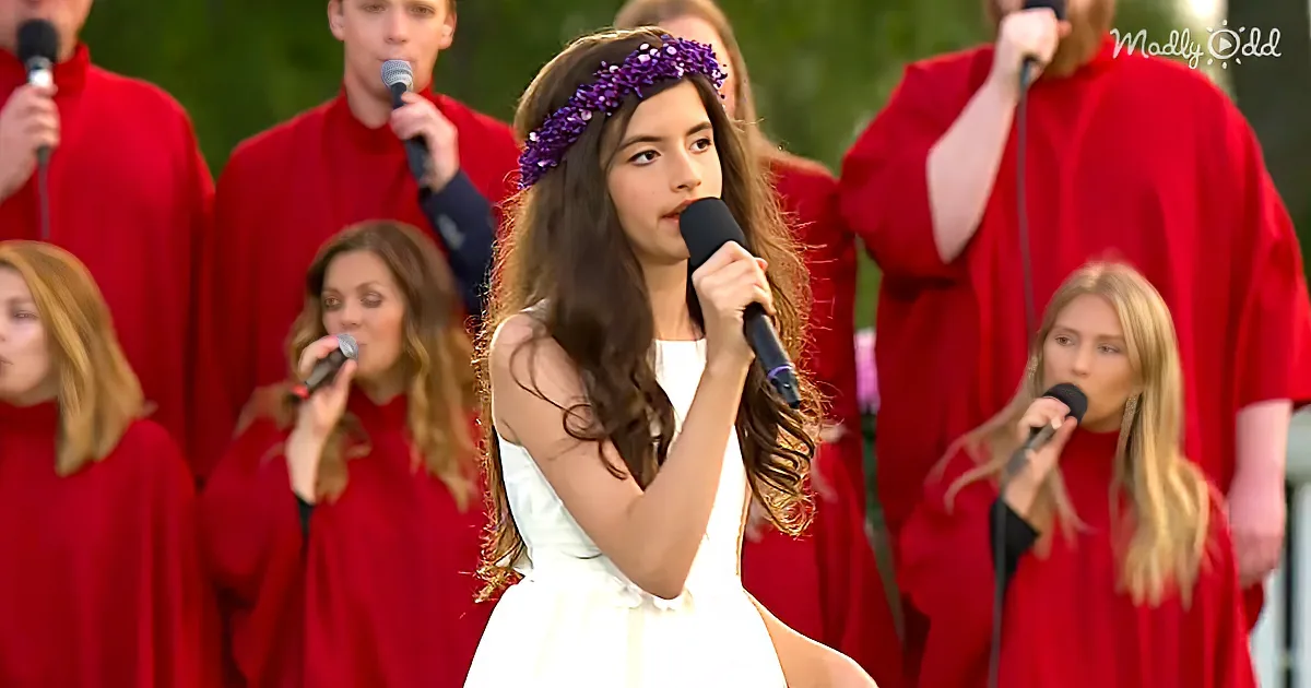 A young woman in white and purple floral crown sings into a microphone, surrounded by a choir in red robes.