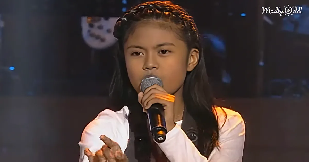 A young girl with braids holds a microphone, wearing a white shirt, and appears to be singing or speaking on stage.