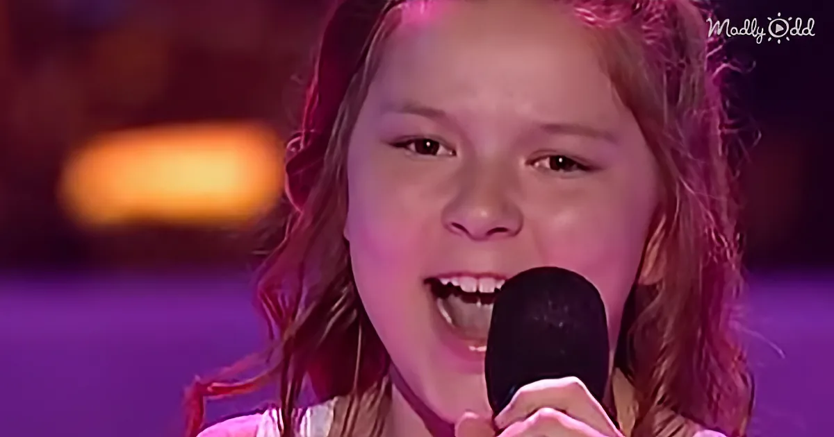 A young girl with long, wavy hair sings into a microphone on a stage with purple lighting.