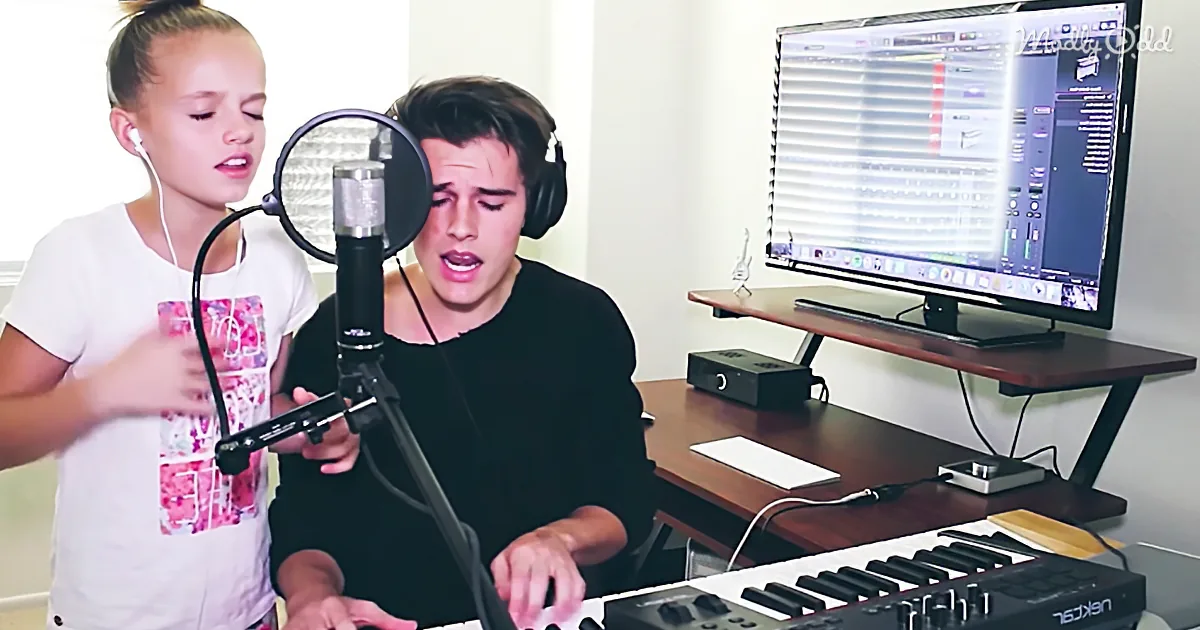A young man sings into a microphone while playing a keyboard, with a young girl standing nearby, both in a home recording...