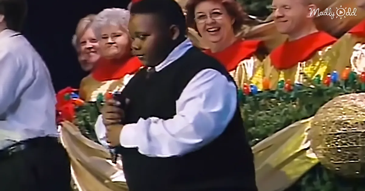 A young boy in a white shirt and black vest holds a microphone, standing in front of a group of people in festive attire.