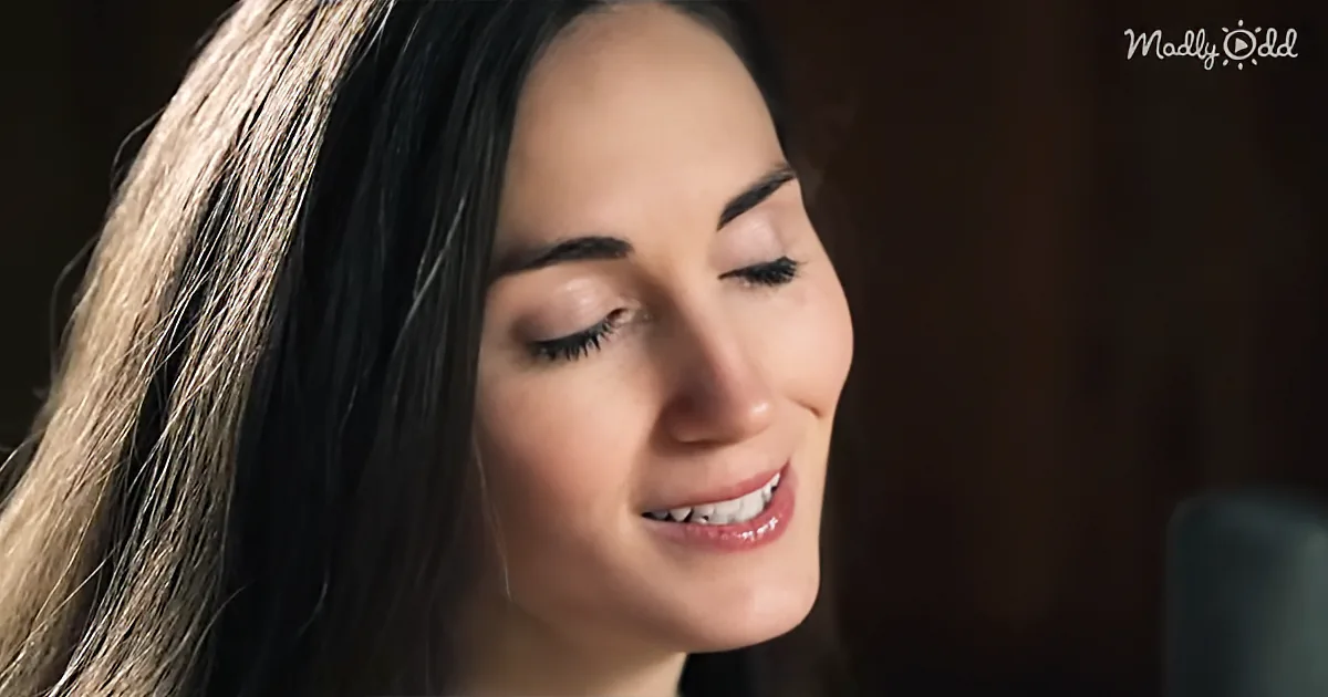 A woman with long dark hair and a smile, looking down, with a microphone in front of her.