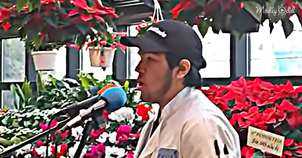 A person wearing a cap sings into a microphone in front of potted red flowers and greenery.