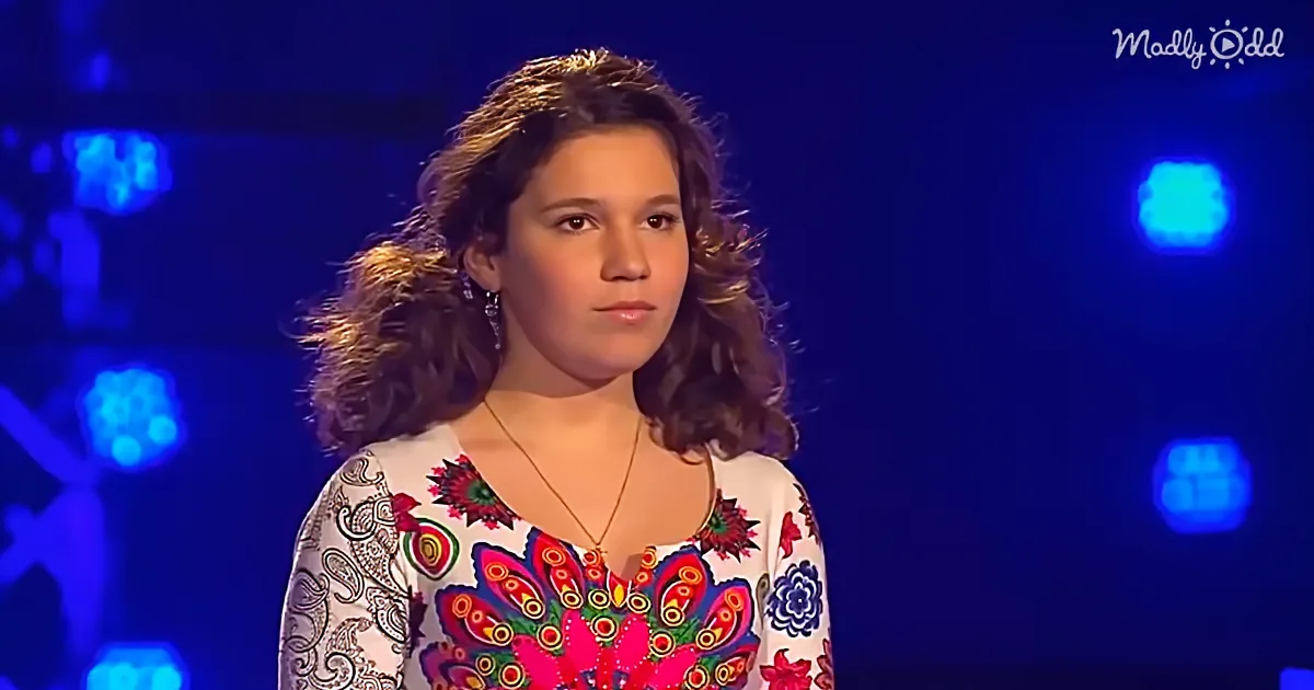 A woman with curly hair, wearing a colorful floral top, stands against a dark background with blue lights.