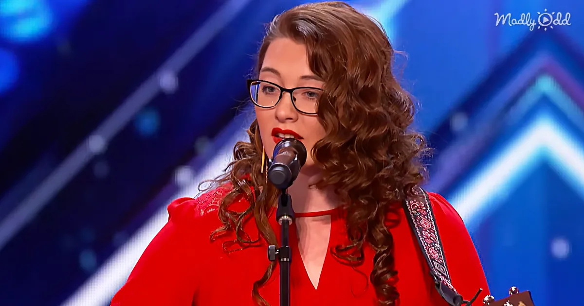 A woman with curly hair and glasses sings into a microphone, wearing a red dress, with a guitar strap visible.
