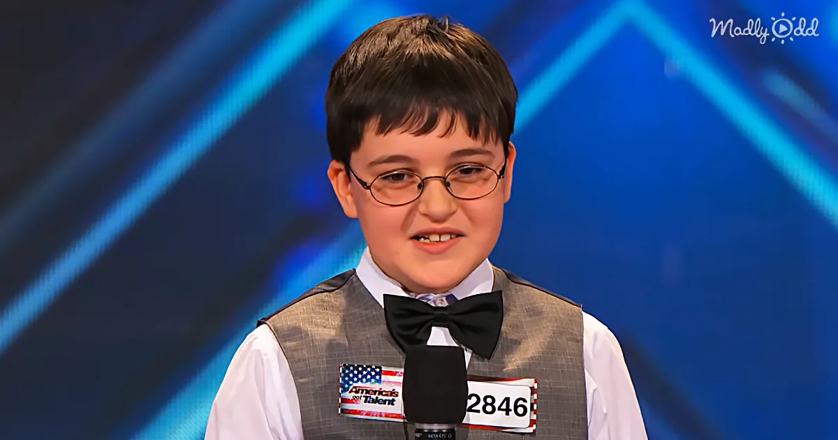 A young boy in a vest and bow tie stands on stage with a microphone, wearing glasses, and a name tag.