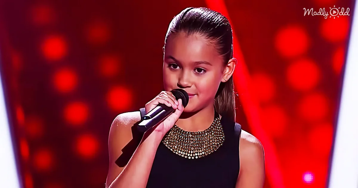 A young girl with a microphone, wearing a black dress and a gold necklace, stands on a stage with a red background.