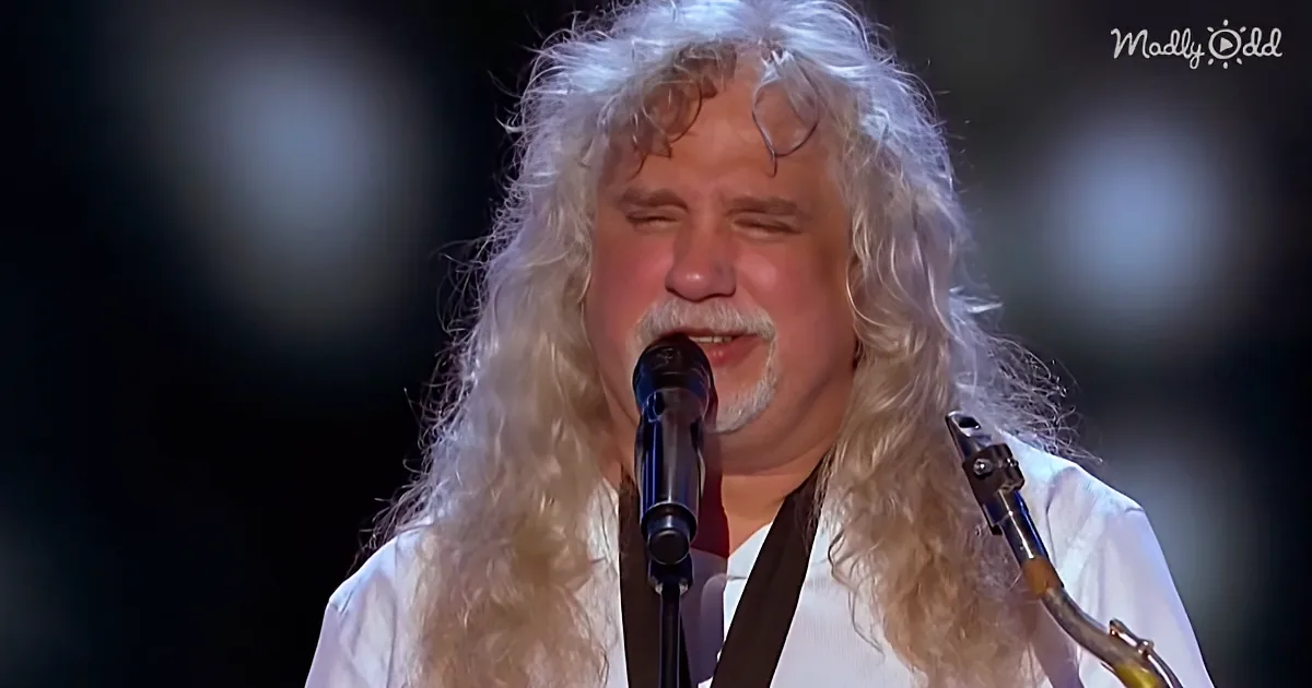 A person with long, curly white hair sings into a microphone, wearing a white shirt.