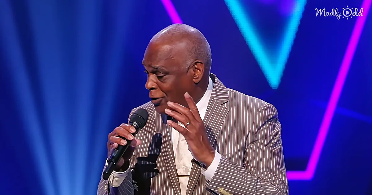 A man in a striped suit speaks into a microphone on a stage with blue and purple lights.