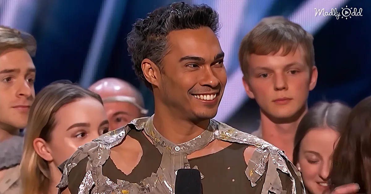 A man in a shiny, metallic outfit smiles while holding a microphone, surrounded by a group of people.