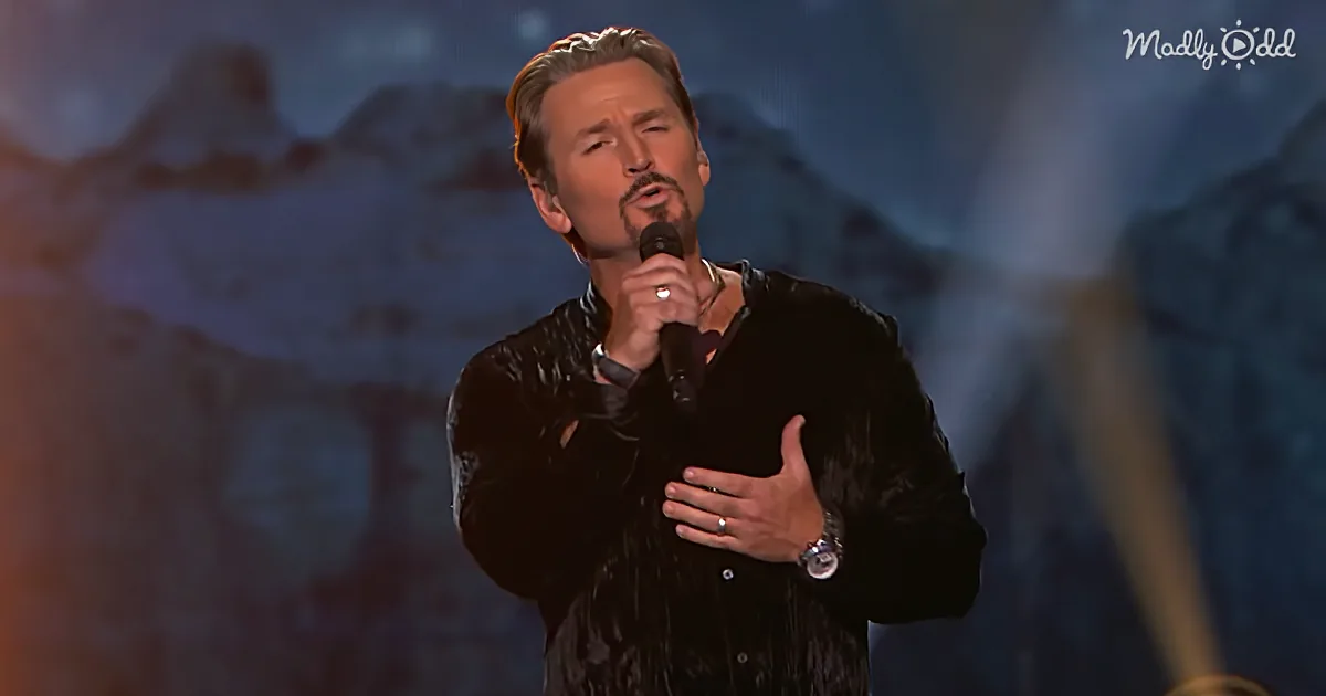 A man in a black jacket sings into a microphone, with a mountainous backdrop.