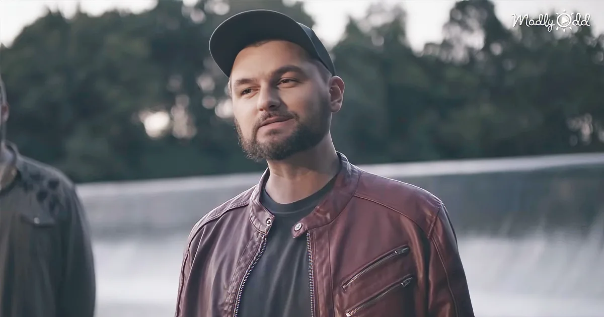 A man in a red leather jacket and cap stands outdoors, looking to the side with a calm expression.