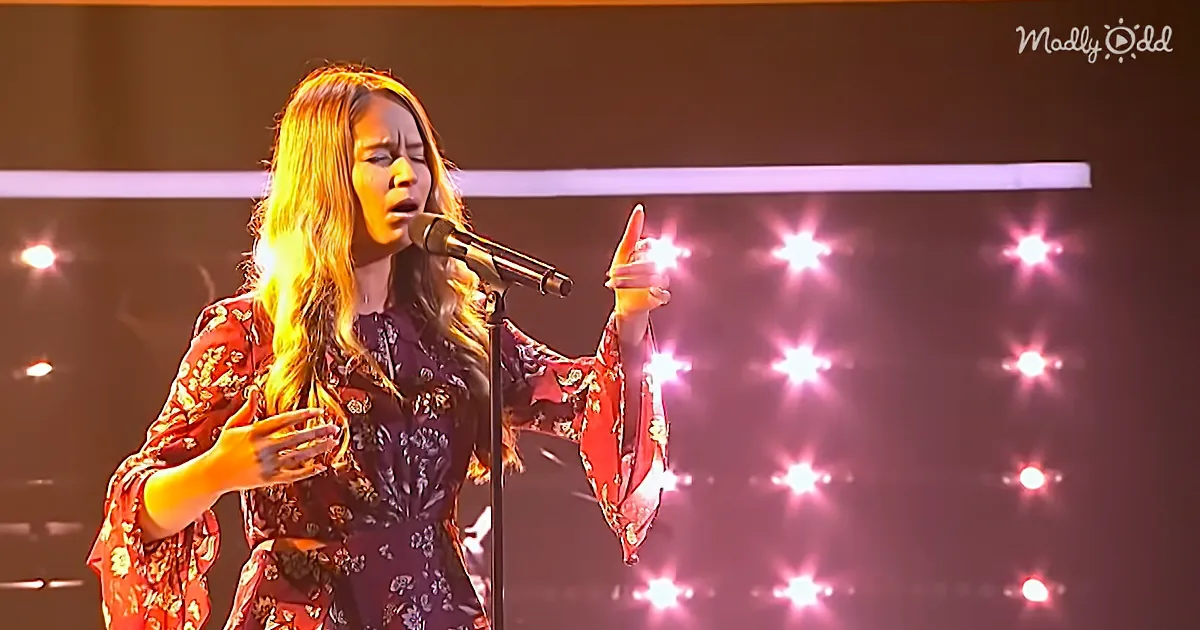 A woman in a floral outfit sings passionately on stage with bright pink lights behind her.