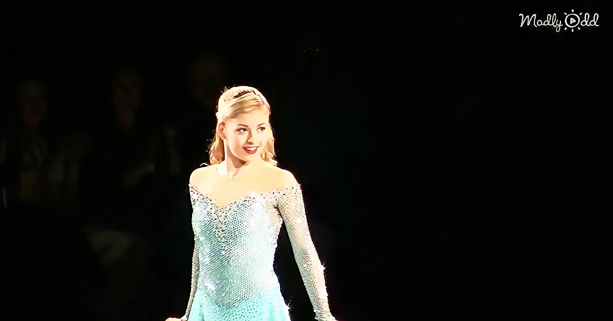 A woman in a sparkling teal dress stands illuminated on stage, smiling.