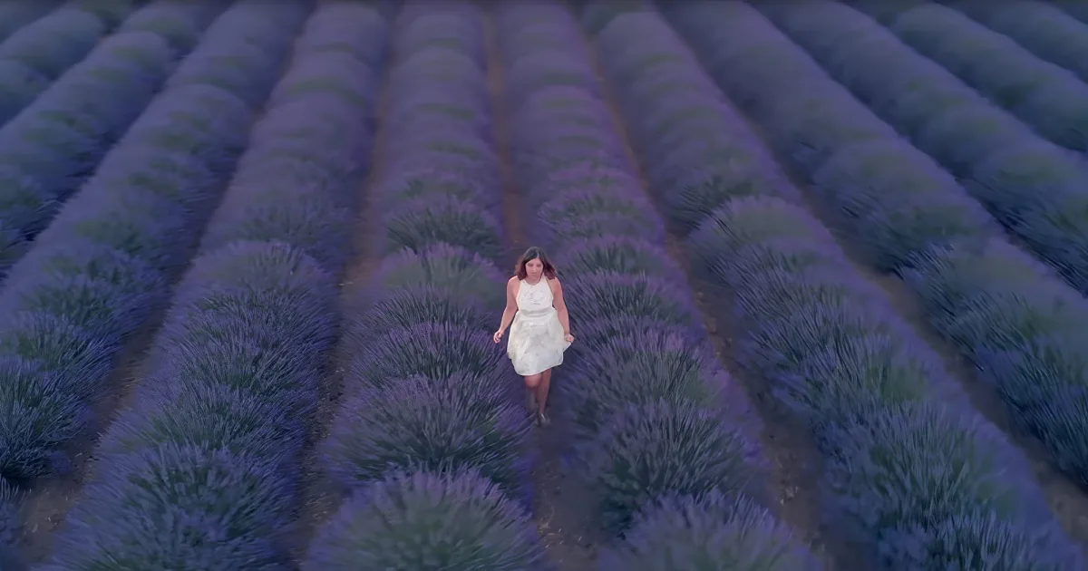 A woman in a white dress walks through a lavender field with rows of purple flowers.