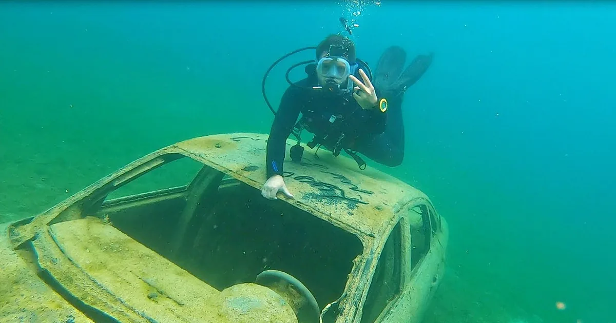 A scuba diver poses on a submerged, rusted car, giving a peace sign underwater.