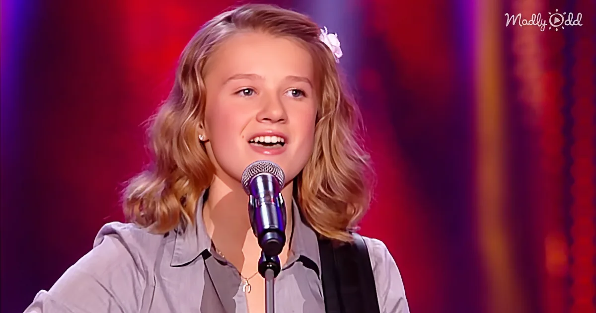 A young girl with blonde hair and a flower in her hair sings into a microphone on stage.
