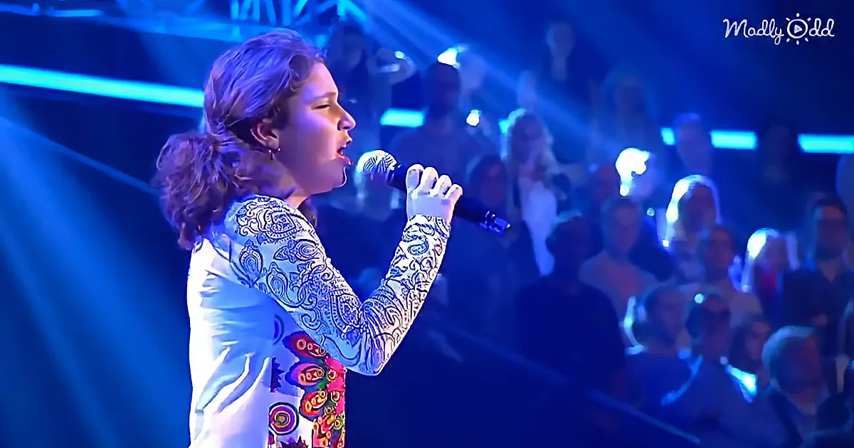 A person sings into a microphone on stage, illuminated by blue lights, with an audience in the background.