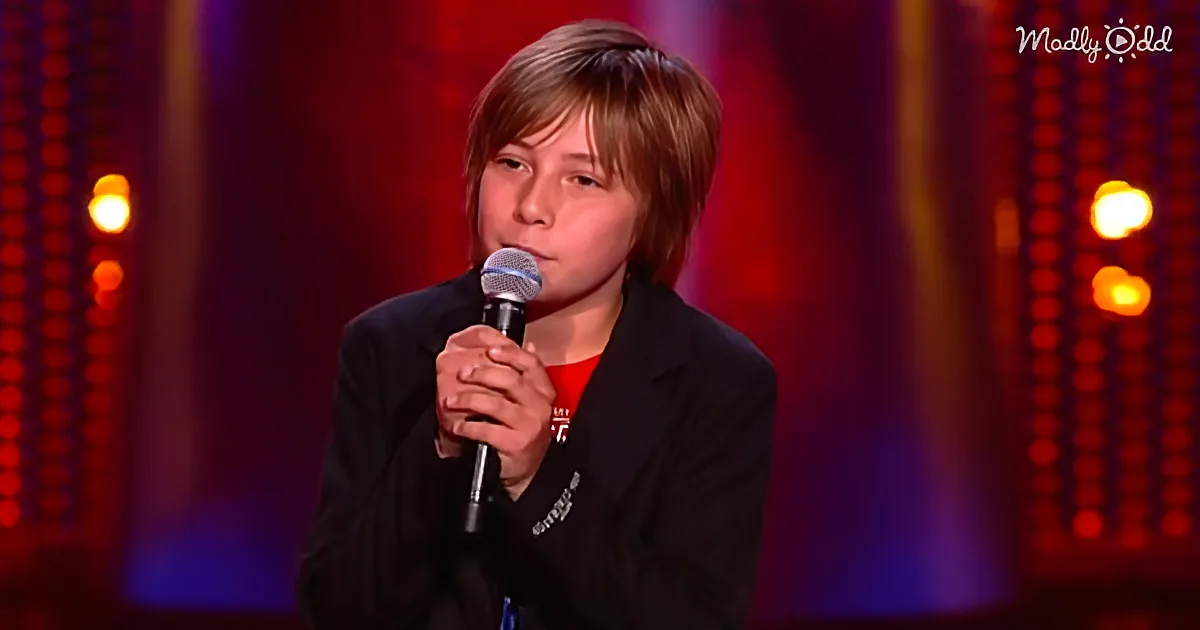 A young person in a black blazer holds a microphone on stage with a red and blue background.