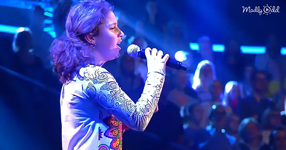 A person with long hair and a patterned shirt sings into a microphone on stage.