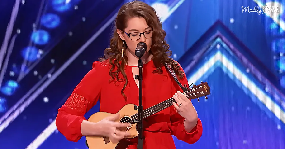 A woman in a red dress plays a ukulele and sings into a microphone on a stage with a blue background.